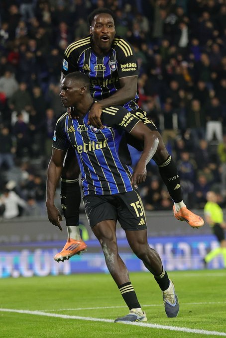 Two Black male soccer players in blue and black striped Atalanta uniforms with yellow accents celebrate on a green soccer field surrounded by a blurred stadium crowd. The player on shoulders wears number 75 jersey and orange cleats, raising arms in joy. The carrying player has muscular build and wears number 17 shorts. They appear ecstatic after scoring.