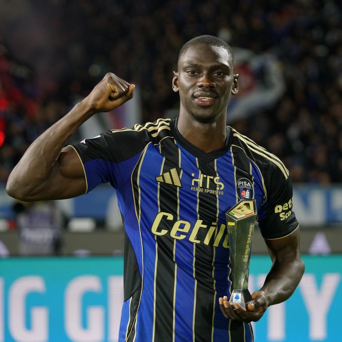 A Black male soccer player with short hair and a smile stands on a green field in a stadium, raising his right fist in celebration. He wears a blue and black striped Adidas jersey with sponsor logos including Celtil and Hts, holding a silver trophy in his left hand. The background shows a crowd of spectators and stadium seating.