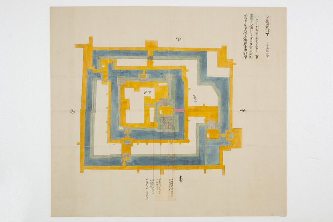 Beige paper with hand-drawn architectural diagram in black ink outlines and yellow blue shading. Central rectangular structure labeled with Japanese text for rooms like bitodokoro bansho daidokoro. Surrounding walls gates paths marked A B C D. Annotations in Japanese script detail fire damage spots from 1849 temple incident at Yonezawa Castle Ninomaru.