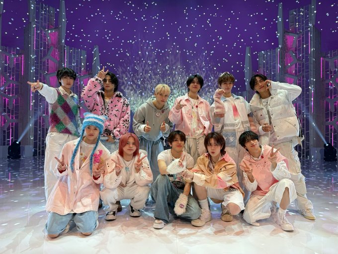 Eleven young men in colorful patterned jackets, sweaters, and pants in pink, white, blue, and purple tones pose energetically on a stage with purple lighting, sparkling confetti, and tall metallic pillar backdrops. Some point fingers, others kneel or stand with hands on hips, all smiling or focused expressions. Outfits include beanies, scarves, and sneakers suggesting a festive performance setting.