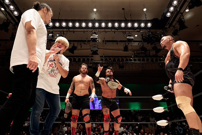 Four male wrestlers in a ring during a professional wrestling event. One with long black hair wears white t-shirt and jeans standing outside ropes gesturing. Another with blonde hair in white t-shirt and black pants stands nearby. Two muscular shirtless wrestlers in black shorts and knee pads pose with arms raised holding championship belts one with tattoos and beard the other bald and larger build. Green lighting and audience in background with overhead lights.