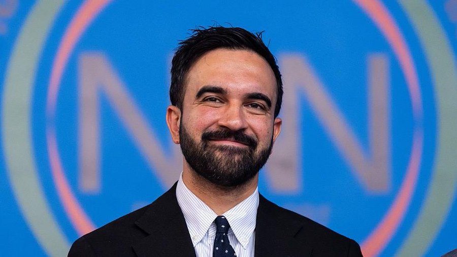 First image shows a man with dark hair and beard smiling while wearing a dark suit white shirt and tie standing in front of a blue background with white N letters. Second image depicts a group of people in blue clothing walking on a stone-paved path near ancient stone walls under a clear sky holding Israeli flags and wearing sunglasses some carrying water bottles.