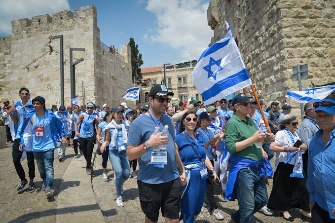 First image shows a man with dark hair and beard smiling while wearing a dark suit white shirt and tie standing in front of a blue background with white N letters. Second image depicts a group of people in blue clothing walking on a stone-paved path near ancient stone walls under a clear sky holding Israeli flags and wearing sunglasses some carrying water bottles.