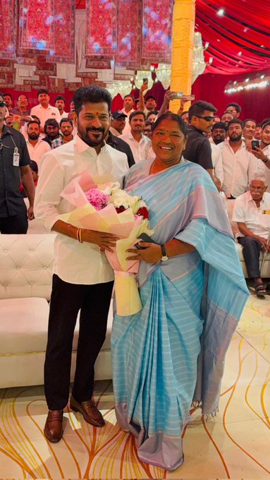 A woman in a light blue saree with a broad smile holds a bouquet of pink roses and white flowers while standing beside a man in a white kurta-pajama with a trimmed beard also smiling, both positioned in front of a white sofa at an indoor event space decorated with red and gold drapes, hanging lights, and a yellow pillar, surrounded by men in traditional attire observing the scene.