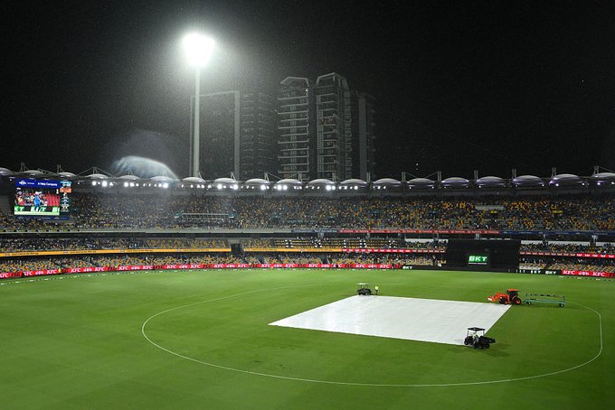 Nighttime view of a large outdoor cricket stadium with green field and white pitch covers being placed by small vehicles. Bright floodlights illuminate the area. Packed yellow seating stands filled with spectators. Large video screen displays BGT. Background features tall modern buildings and advertising banners around the boundary.