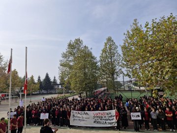 First image shows a large group of students mostly in red attire gathered on a school sports field holding banners reading YASIMA VATAN and ATATÜRKE with Turkish flags in the background and trees around. Second image depicts a similar crowd of students in red clothing on the field with banners stating YASIMA VATAN and ATANURKE MINNETLE ANIYORUZ standing near a podium under clear skies with flags and fences. Third image features five students in school uniforms including red polos kneeling or standing around a small newly planted sapling on grassy ground using a shovel with a sign placed nearby. Fourth image captures a group of students and adults including some in red shirts planting a small tree on grass with a sign holding tools like a shovel under trees and a flag in the background.