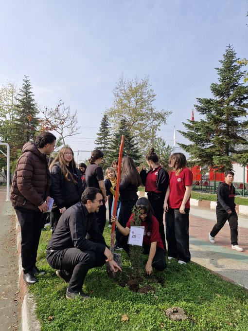 First image shows a large group of students mostly in red attire gathered on a school sports field holding banners reading YASIMA VATAN and ATATÜRKE with Turkish flags in the background and trees around. Second image depicts a similar crowd of students in red clothing on the field with banners stating YASIMA VATAN and ATANURKE MINNETLE ANIYORUZ standing near a podium under clear skies with flags and fences. Third image features five students in school uniforms including red polos kneeling or standing around a small newly planted sapling on grassy ground using a shovel with a sign placed nearby. Fourth image captures a group of students and adults including some in red shirts planting a small tree on grass with a sign holding tools like a shovel under trees and a flag in the background.