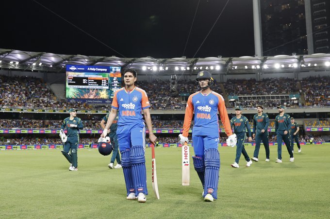 Two Indian cricket players in blue uniforms with orange accents stand on a green field holding bats one with a helmet under arm during night match at stadium with bright lights and large electronic scoreboard displaying scores behind them several Australian players in green uniforms walk in background crowd visible in stands BCCI logo on bats and jerseys