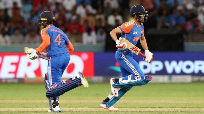 Two male cricketers in blue India national team uniforms with orange accents and number 4 on one jersey run on a green grass field during a match with a blurred stadium crowd in the background and advertising banners visible including one for DP World.