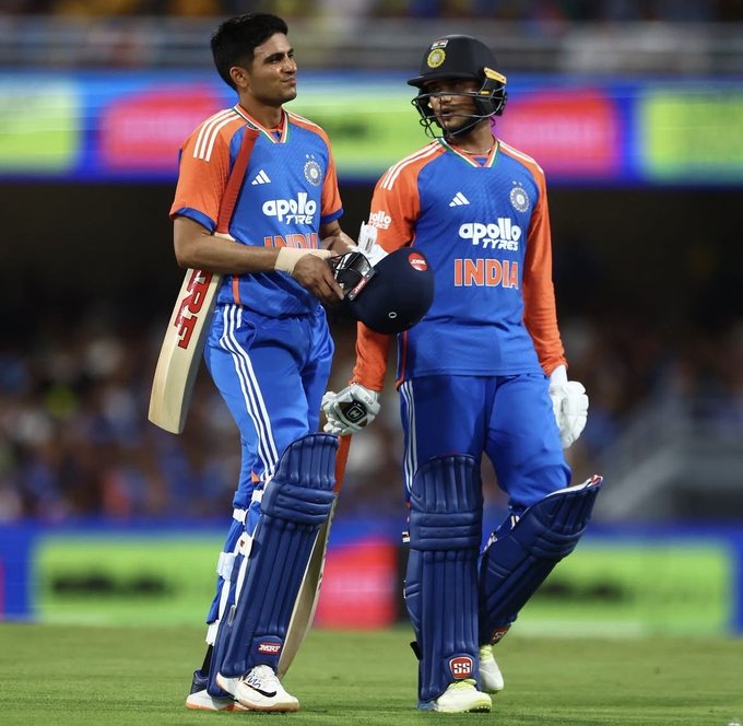 Two male cricketers in blue Team India uniforms with orange accents stand side by side on a green cricket field during a match. The player on the left holds a bat over his shoulder and wears leg pads and gloves. The player on the right holds a helmet in one hand and wears batting pads and gloves. Both have protective gear and the background shows stadium seating with orange and blue sponsor logos like Apollo Tyres.