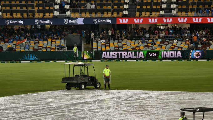 A cricket stadium filled with spectators in yellow and orange seats under a partly cloudy sky. The field is covered with white tarps to protect against rain, with a golf cart and ground staff nearby. Large banners display Australia vs India and sponsor logos like Remitly. The scoreboard and seating areas are visible in the background.