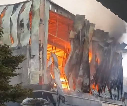 A multi-story industrial building engulfed in flames with intense orange fire visible through broken windows and on the roof. Smoke billows from the structure amid a daytime scene with some greenery nearby. The facade shows damage from the blaze including charred edges and exposed framework.