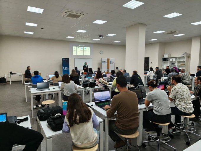 First image shows a spacious classroom with fluorescent ceiling lights, white walls, and pillars. Multiple rows of tables with participants seated, using laptops and notebooks. Some stand near a projector screen displaying charts. A banner with BTK Akademi logo is visible on the wall. Diverse group of adults and young people in casual attire engage in training activities. Second image depicts a similar classroom setup with a projector screen showing BTK Akademi logo. Participants sit at tables with laptops, some wearing headscarves. Instructors stand near a whiteboard and banners for BTK and collaborators. Clock on wall indicates time, and group appears focused on session.