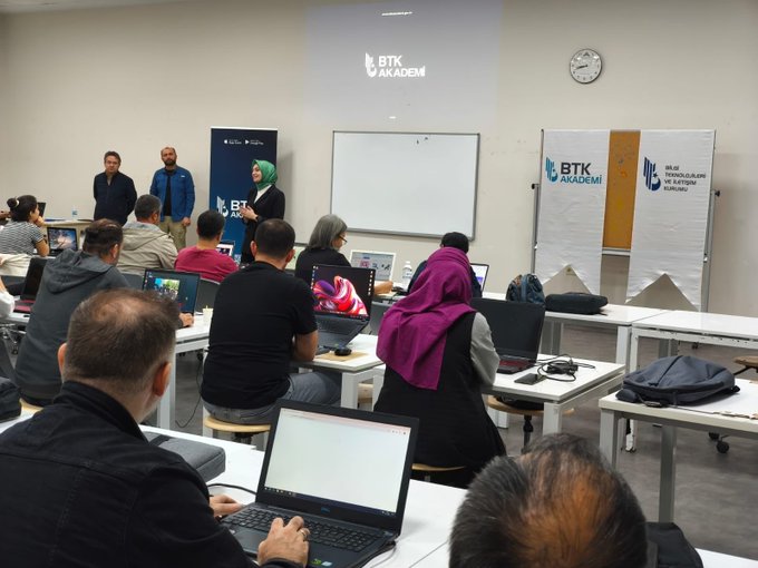 First image shows a spacious classroom with fluorescent ceiling lights, white walls, and pillars. Multiple rows of tables with participants seated, using laptops and notebooks. Some stand near a projector screen displaying charts. A banner with BTK Akademi logo is visible on the wall. Diverse group of adults and young people in casual attire engage in training activities. Second image depicts a similar classroom setup with a projector screen showing BTK Akademi logo. Participants sit at tables with laptops, some wearing headscarves. Instructors stand near a whiteboard and banners for BTK and collaborators. Clock on wall indicates time, and group appears focused on session.