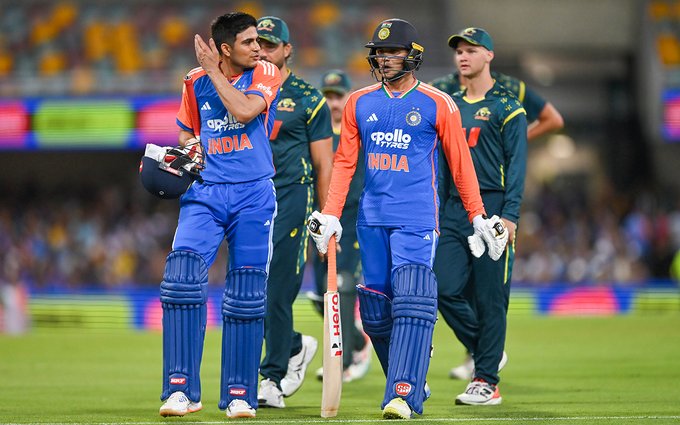 Indian cricket players Shubman Gill and Abhishek Sharma in blue uniforms with orange accents stand on the field wearing helmets and gloves holding a bat interacting with Australian players in green uniforms at the Gabba stadium during the match surrounded by spectators and advertising boards.