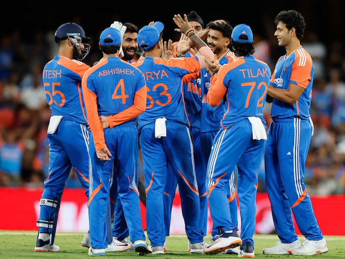 Group of Indian cricket players in blue jerseys with numbers like 55 on backs including names Rishabh Pant Tilak Varma Abhishek Sharma celebrating by raising hands and smiling on green field with blurred stadium crowd in background under overcast sky