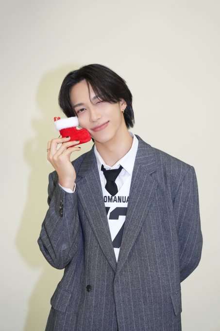Young man with black hair in a gray pinstripe suit white shirt black tie stands against a light background holding a small red object near his smiling face with hand near mouth in playful pose.