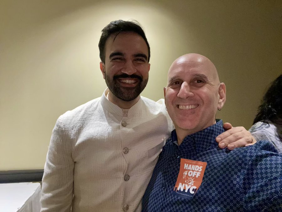 Two men stand side by side in a warmly lit indoor setting with beige walls. The man on the left wears a white kurta with buttons and smiles broadly showing teeth. The man on the right is bald wears a blue collared shirt with an orange badge labeled Hands Off NYC and has his arm around the other mans shoulder both smiling. A woman with dark hair is partially visible in the background.