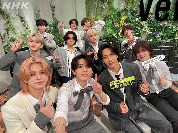 Group of eleven young men from INI posing together in a studio setting with green plants and flowers in background wearing formal suits shirts and ties some with jackets holding signs that say Venue101 and NHK ONE making peace signs and smiling at camera.