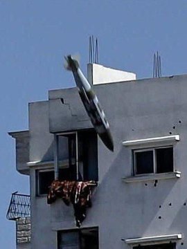 First image shows a white building with beige stone base, multiple windows some with bars and laundry hanging from one balcony, bullet holes in walls, and a gray cylindrical bomb or missile angled upward near the top right corner against a clear blue sky with construction rebar visible. Second image depicts a large plume of thick gray smoke rising from the ground near several damaged beige buildings with debris, surrounded by scattered white tents and makeshift shelters in an open area under a clear blue sky.