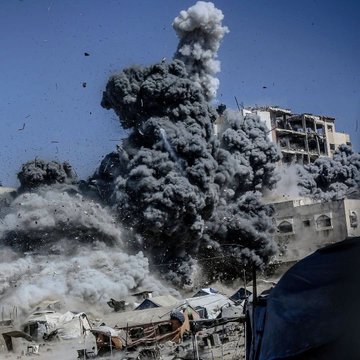 First image shows a white building with beige stone base, multiple windows some with bars and laundry hanging from one balcony, bullet holes in walls, and a gray cylindrical bomb or missile angled upward near the top right corner against a clear blue sky with construction rebar visible. Second image depicts a large plume of thick gray smoke rising from the ground near several damaged beige buildings with debris, surrounded by scattered white tents and makeshift shelters in an open area under a clear blue sky.
