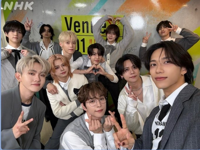 Eleven young men in coordinated white and gray outfits with accessories pose energetically in front of a yellow and orange Venue101 banner on a wall, some making peace signs, heart gestures, and smiles, with the group name INI implied by context.