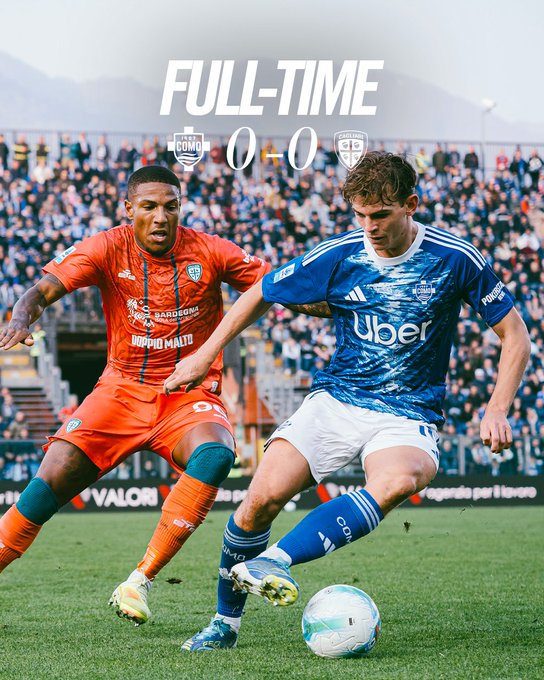 Two soccer players compete for a white ball on green field turf under stadium lights with crowd in background. Left player in orange jersey with black accents and tattoos on arms wears orange shorts and yellow-green cleats. Right player in blue jersey with white patterns and Uber logo wears blue shorts and blue-white cleats. Full-time 0-0 score overlay in white text at top.