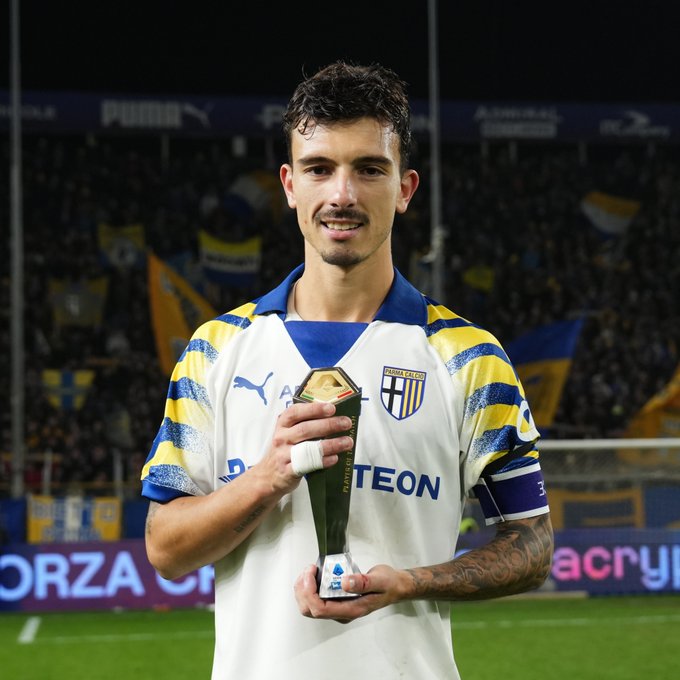 A male athlete with curly dark hair and tattoos on his arms stands on a soccer field holding a trophy with both hands. He wears a yellow and blue striped Puma jersey with the Atalanta logo. The background shows a stadium with blurred crowd and green grass. Sponsor logos like Teon and Zacry appear on banners.