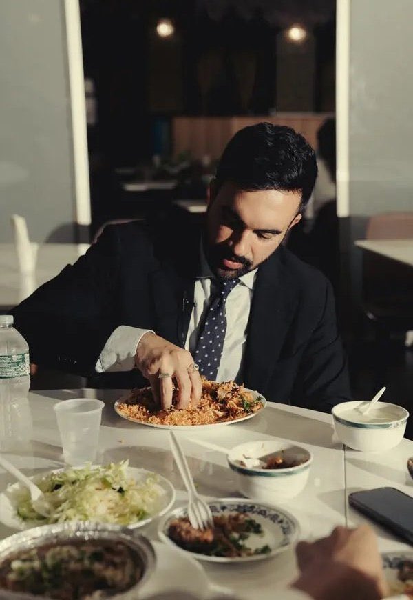 A man with short dark hair wearing a dark suit, white shirt, and striped tie sits at a round white table in a softly lit indoor space with beige walls and hanging lights. He uses his right hand to scoop rice from a white plate filled with yellow rice and small vegetables. Nearby plates include a white dish with green lettuce salad and chopsticks, a small blue-rimmed bowl with brownish soup and spoon, a foil takeout container with reddish-brown saucy food, and another small bowl with greens. A clear plastic cup with lid and a water bottle are on the table, along with a black smartphone. Another hand reaches in from the side holding chopsticks near a plate.