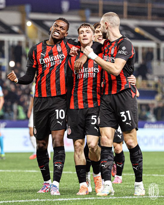 Four male soccer players wearing red and black striped AC Milan jerseys with sponsor logos stand on a green field in a stadium under bright lights. They embrace each other with arms around shoulders, smiling and appearing joyful. One player has dreadlocks, another short blond hair, and they wear matching shorts, socks, and cleats. Stadium seating and advertising boards are visible in the background.