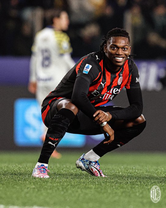 Rafael Leao crouches on green grass field in stadium, smiling with dreadlocks, wearing red and black AC Milan striped jersey with sponsor logos, black shorts and sleeves, knee pads, colorful patterned cleats, ring on finger, another player in white and yellow kit blurred in background, purple stadium seating and crowd visible.