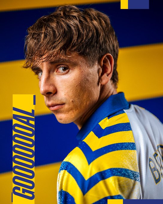 A young male soccer player with brown hair and focused expression stands in profile view, wearing a white and blue striped jersey with yellow accents, positioned against a background of vertical yellow and blue stripes, with overlaid text GOAL in large yellow letters on a blue banner.