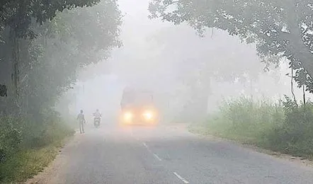 A rural road lined with trees and grass shrouded in thick fog with a bus driving on the left side its headlights on and visible through the mist a motorcycle with rider behind it and two distant figures walking on the right side of the road.