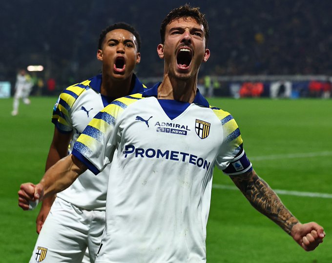 Two male soccer players wearing yellow and blue striped Parma Calcio jerseys with sponsor logos stand on a green soccer field at night under stadium lights one player in foreground has tattoos on arms and raises both arms in celebration shouting with open mouth the other player behind him also shouts excitedly with open mouth both wear matching shorts and socks crowd blurred in background