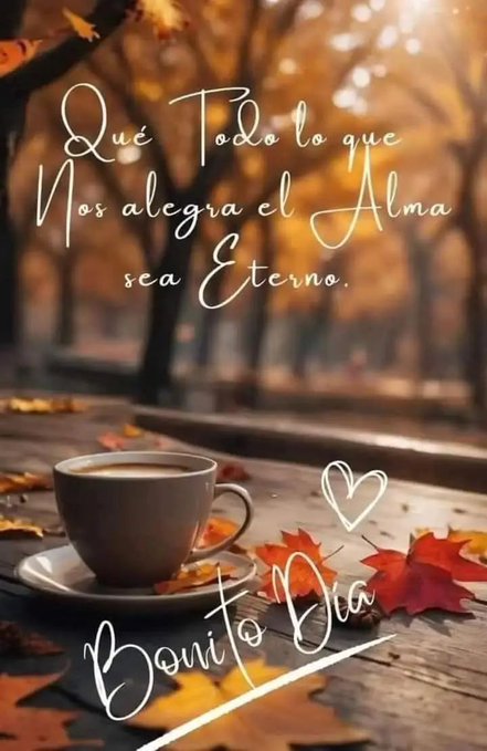 A close-up view of a rustic wooden table surface with a white saucer holding a light brown ceramic cup filled with coffee, steam subtly rising from it. Several vibrant red and orange autumn leaves are scattered around the cup and on the saucer. In the blurred background, a forest scene with trees bearing autumn foliage in shades of yellow and orange under soft golden sunlight filtering through branches. Overlaid text in elegant cursive script reads Que Todos lo que alma nos alegra Nos alegrará el Alma sea Eterno, with a pink heart symbol and Bonito Día below.