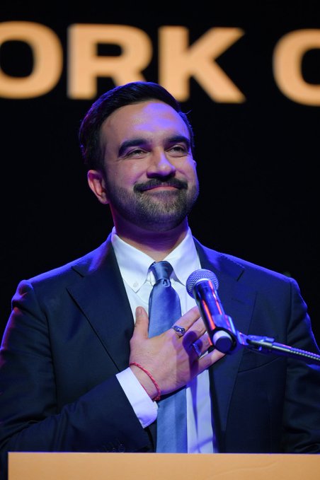 First image shows a man with short dark hair and a mustache wearing a dark suit white shirt blue tie and red wristband holding a microphone in his right hand while standing at a wooden podium with a black background featuring illuminated text WORKORG in white letters. Second image depicts an elderly man with white hair wearing a dark suit white shirt blue tie and an American flag pin standing at a wooden podium gesturing with his right hand surrounded by multiple blue and white Israeli flags in a formal setting.