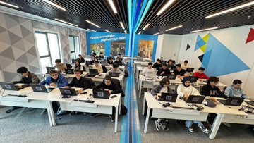 First image shows a brightly lit classroom with rows of desks where young students in casual clothing sit facing laptops, some wearing hoodies and backpacks, with a teacher standing nearby pointing at a large screen displaying code or slides on the wall, modern architecture with large windows and beige walls. Second image depicts another classroom setup with students at long white tables using laptops, a screen showing a brain diagram or network graphic, participants in varied attire including jackets and scarves, high ceiling with exposed beams and glass walls overlooking an outdoor area. Third image features two rows of desks in a colorful room with blue and geometric wall designs, students focused on laptops, some with notebooks, wearing sneakers and casual outfits, a banner reading something in Turkish on the wall. Fourth image displays a large group of young adults and students posing together in a conference-like room with a dark wall featuring the BTK Akademi logo, cloud icons, and Turkish text, individuals in professional and casual clothes smiling at the camera, standing on a gray floor.