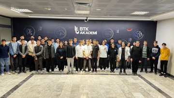 First image shows a brightly lit classroom with rows of desks where young students in casual clothing sit facing laptops, some wearing hoodies and backpacks, with a teacher standing nearby pointing at a large screen displaying code or slides on the wall, modern architecture with large windows and beige walls. Second image depicts another classroom setup with students at long white tables using laptops, a screen showing a brain diagram or network graphic, participants in varied attire including jackets and scarves, high ceiling with exposed beams and glass walls overlooking an outdoor area. Third image features two rows of desks in a colorful room with blue and geometric wall designs, students focused on laptops, some with notebooks, wearing sneakers and casual outfits, a banner reading something in Turkish on the wall. Fourth image displays a large group of young adults and students posing together in a conference-like room with a dark wall featuring the BTK Akademi logo, cloud icons, and Turkish text, individuals in professional and casual clothes smiling at the camera, standing on a gray floor.