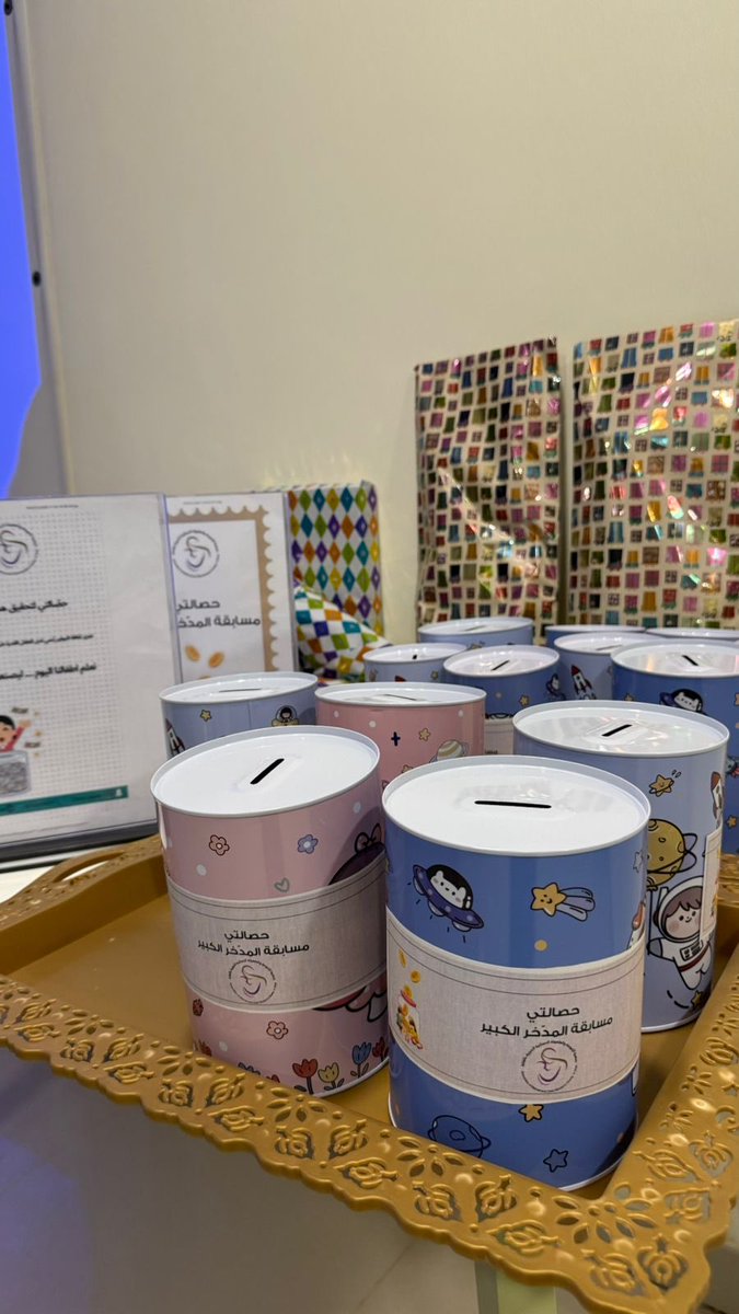 First image shows group of young children mostly girls in colorful casual clothes sitting on floor in indoor room with beige walls plants and chairs some smiling others looking at camera wearing headbands. Second image displays multiple pink and blue piggy banks with decorative patterns on tray next to wrapped gifts and certificate frames in light room. Third image features projector screen with Arabic text about financial awareness and hand putting coin in jar illustration surrounded by stacked colorful wrapped gifts and educational posters. Fourth image projects banner with charity logo Arabic text on financial awareness for children showing hands inserting coins into jars on wall in conference-like setting.