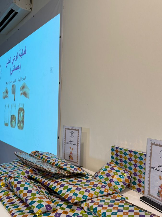 First image shows group of young children mostly girls in colorful casual clothes sitting on floor in indoor room with beige walls plants and chairs some smiling others looking at camera wearing headbands. Second image displays multiple pink and blue piggy banks with decorative patterns on tray next to wrapped gifts and certificate frames in light room. Third image features projector screen with Arabic text about financial awareness and hand putting coin in jar illustration surrounded by stacked colorful wrapped gifts and educational posters. Fourth image projects banner with charity logo Arabic text on financial awareness for children showing hands inserting coins into jars on wall in conference-like setting.