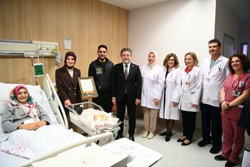 First image shows a man in a dark suit holding a newborn baby wrapped in a white blanket with embroidery in a hospital room, smiling while surrounded by medical staff in white coats and scrubs standing nearby. Second image depicts a group including a suited man, a woman in a headscarf holding a baby, and others presenting a framed certificate in a decorated hospital room with pink balloons and baby items. Third image features a larger group of hospital staff and officials, some in headscarves and lab coats, gathered around a crib with a baby in a hospital bed. Fourth image shows another group of people including suited individuals and medical personnel posing near a hospital bed with a crib containing a baby.