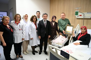 First image shows a man in a dark suit holding a newborn baby wrapped in a white blanket with embroidery in a hospital room, smiling while surrounded by medical staff in white coats and scrubs standing nearby. Second image depicts a group including a suited man, a woman in a headscarf holding a baby, and others presenting a framed certificate in a decorated hospital room with pink balloons and baby items. Third image features a larger group of hospital staff and officials, some in headscarves and lab coats, gathered around a crib with a baby in a hospital bed. Fourth image shows another group of people including suited individuals and medical personnel posing near a hospital bed with a crib containing a baby.