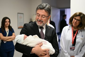 First image shows a man in a dark suit holding a newborn baby wrapped in a white blanket with embroidery in a hospital room, smiling while surrounded by medical staff in white coats and scrubs standing nearby. Second image depicts a group including a suited man, a woman in a headscarf holding a baby, and others presenting a framed certificate in a decorated hospital room with pink balloons and baby items. Third image features a larger group of hospital staff and officials, some in headscarves and lab coats, gathered around a crib with a baby in a hospital bed. Fourth image shows another group of people including suited individuals and medical personnel posing near a hospital bed with a crib containing a baby.