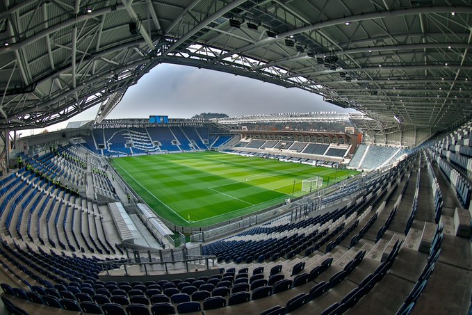 Empty football stadium viewed from stands showing green grass field with white goalposts, blue and white tiered seating sections, metallic roof trusses overhead, and overcast sky above.