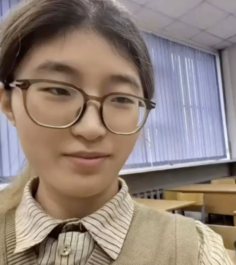 一位棕色直发扎成马尾辫的年轻亚洲女性戴着棕色框框眼镜，米色背心下穿着浅米色衬衫，带有条纹领子。她站在教室里，背景是木桌椅，大窗户上挂着浅蓝色窗帘。