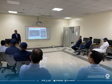First image shows a classroom with men in traditional white thobes and ghutras seated at desks with computers, one man in suit standing and gesturing towards a projector screen displaying software interface, wooden table with equipment, windows with blinds. Second image depicts a trainer in suit presenting to seated participants in suits and caps around computers, projector showing screen content, room with doors and chairs. Third image features group of men in suits and thobes sitting around computers in a spacious room with rainbow flag poster on wall, projector hanging from ceiling. Fourth image illustrates participants in various attire including caps and thobes seated at computers, female in abaya at desk, projector screen visible, room with artwork on walls.