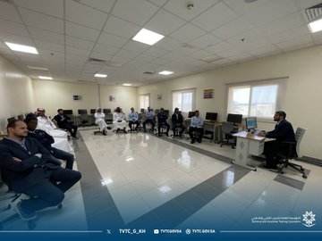 First image shows a classroom with men in traditional white thobes and ghutras seated at desks with computers, one man in suit standing and gesturing towards a projector screen displaying software interface, wooden table with equipment, windows with blinds. Second image depicts a trainer in suit presenting to seated participants in suits and caps around computers, projector showing screen content, room with doors and chairs. Third image features group of men in suits and thobes sitting around computers in a spacious room with rainbow flag poster on wall, projector hanging from ceiling. Fourth image illustrates participants in various attire including caps and thobes seated at computers, female in abaya at desk, projector screen visible, room with artwork on walls.