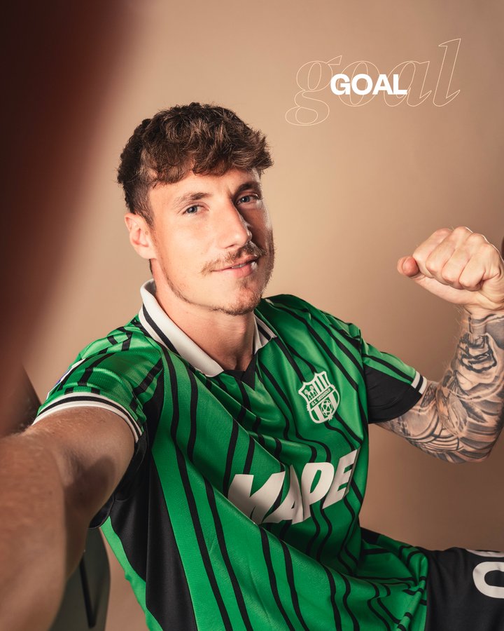 A young man with curly brown hair and a mustache poses confidently in a green and black striped soccer jersey with the Sassuolo logo on the chest and Mapei sponsor text, flexing his right arm while seated, showing tattoos on his arms, against a warm beige background with faint GOAL text overlay.