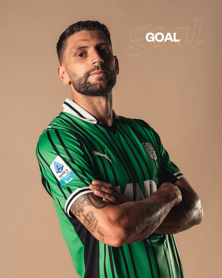 Man with short dark hair and beard wearing green and black striped Puma soccer jersey with white accents and team logo on chest, arms crossed, standing against beige background.