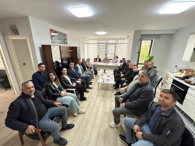 First image displays a group of eight adults seated around an elliptical white table in a modern conference room with large windows covered by sheer blinds allowing natural light, several Turkish flags positioned on the table, participants dressed in formal attire including suits jackets and one headscarf, some holding documents or cups, small plates with food items like pastries on the table, wooden flooring and neutral walls. Second image shows a larger group of about fifteen adults arranged around a rectangular white table in a kitchen-like meeting area with overhead lighting, wooden cabinets and a plant in the background, participants in casual professional clothing such as jackets and jeans, some seated on chairs others standing, plates with snacks on the table, open windows with blinds.